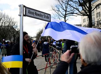 Протестующий с бело-сине-белым флагом стоит под макетом уличного знака с надписью «Улица Алексея Навального» возле посольства России в Берлине.